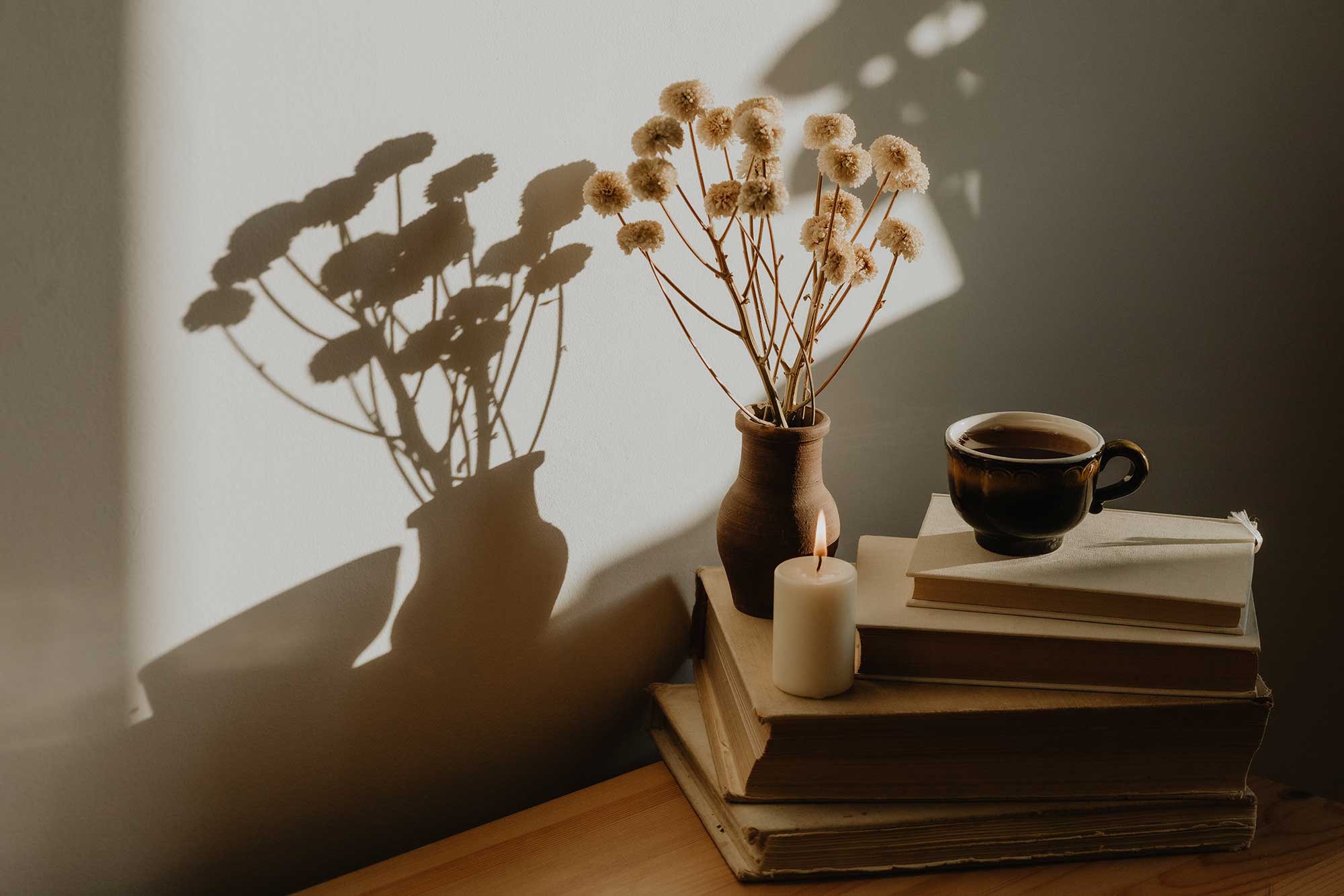 Stillleben mit getrockneten Blumen in einer Vase, einer brennenden Kerze, gestapelten Büchern und einer Tasse auf einem Tisch, mit Schatten an der Wand.