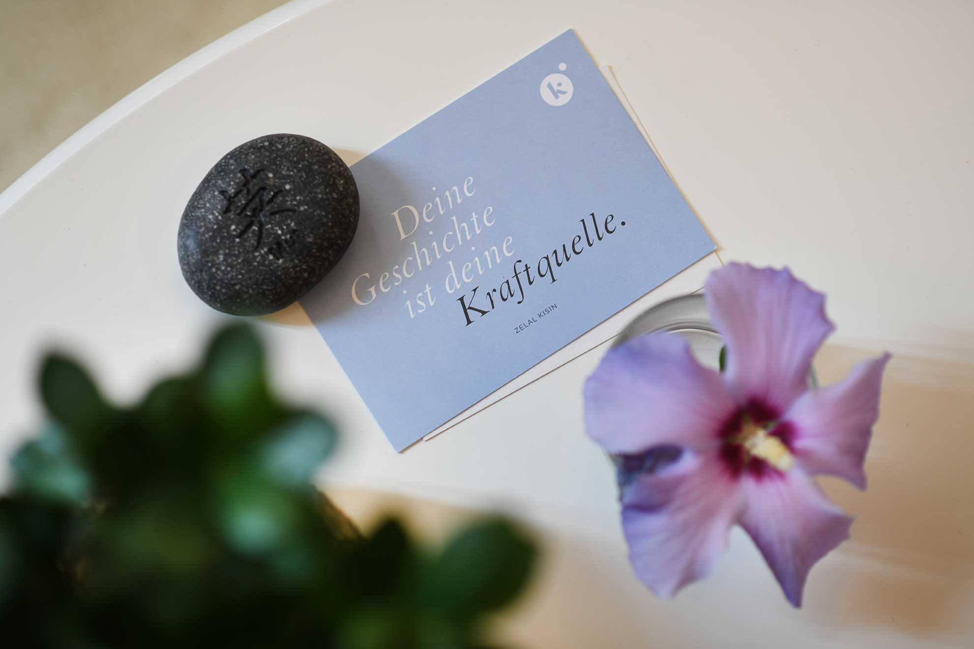 Karte mit dem Schriftzug „Deine Geschichte ist deine Kraftquelle“ liegt auf einem Tisch neben einem dunklen Stein und einer Blüte.